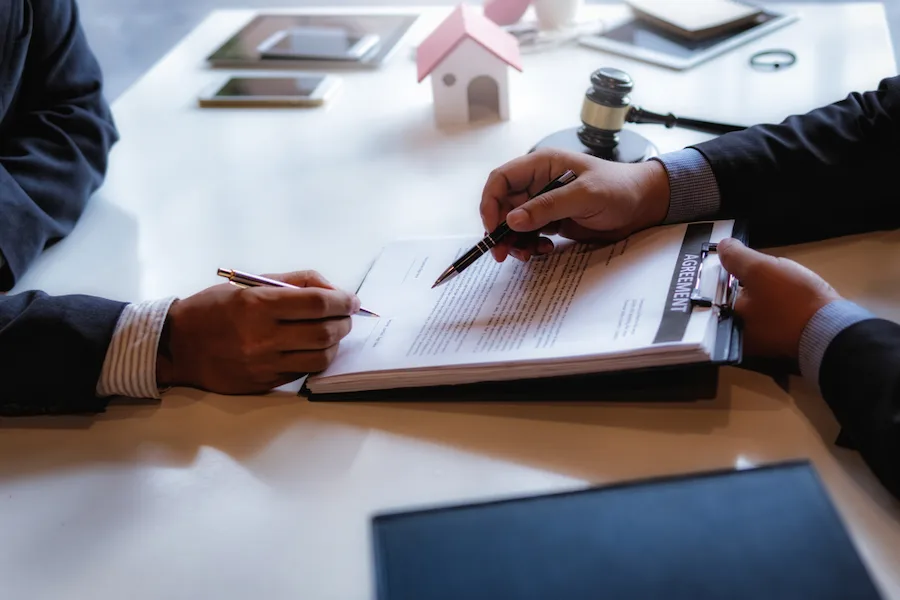 Dos personas revisando y firmando un contrato bancario sobre una mesa, con documentos legales y un mazo judicial al fondo